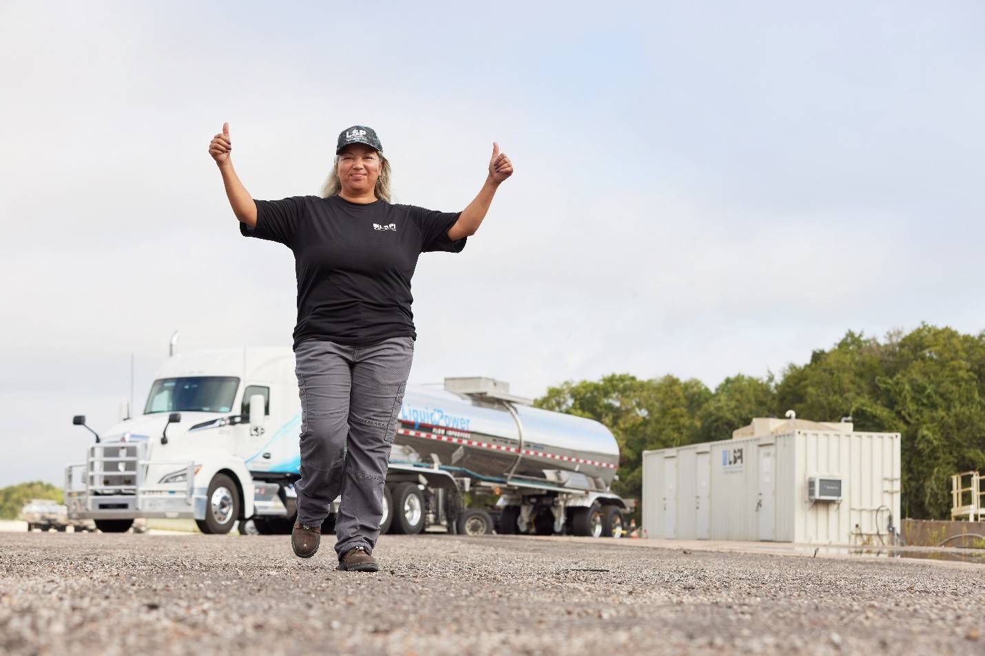 A female commercial professional semi-truck driver is walking towards the camera with both arms up above her head and she’s giving a thumbs up with each hand. In the background there is a truck with a LSP Transport LLC logo on the tractor and a LiquidPower logo on the trailer. You also see a container box skid with the logo LSPI on it.